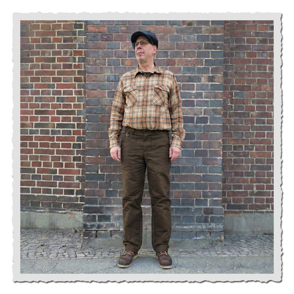 Mann trägt Retronia Arbeiterhose Rudolph II aus braunem Moleskin mit hoher Leibhöhe und geradem Bein im klassischen Workwear-Stil vor Ziegelwand.